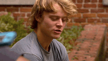 a young man with blonde hair and a gray shirt is sitting in front of a brick wall with his eyes closed .