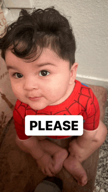 a baby in a spiderman outfit is sitting on the floor with a sign that says please