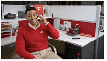 a man in a red sweater is sitting in an office cubicle talking on a phone