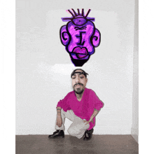 a man in a pink shirt is squatting down in front of a purple drawing on the wall