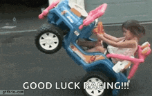 a little girl is sitting in a blue toy car with the words `` good luck racing '' written on it .