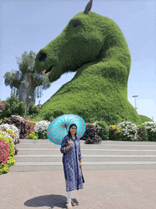 a woman holding a blue umbrella stands in front of a large green horse sculpture