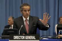 a man in a suit and pink tie stands behind a sign that says director general