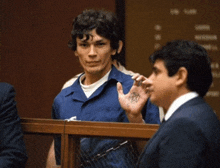 a man in a blue shirt stands in a courtroom talking to a man in a suit