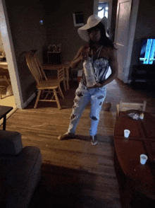 a woman standing in a living room wearing a white hat and ripped jeans