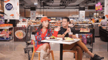 a man and woman sit at a table in a grocery store with a sign that says play growing in the background