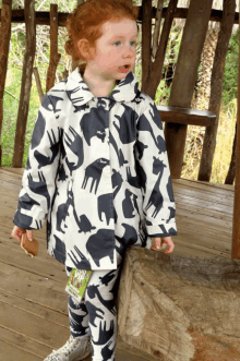 a little girl wearing a black and white coat with animals on it is standing on a wooden deck