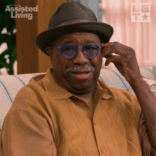 a man wearing glasses and a hat is sitting on a couch with assisted living written on the wall behind him