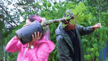 a woman in a pink raincoat is covering her face with a potted plant while a man holds a tree branch over her head