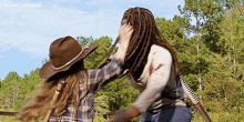 two women with dreadlocks are standing next to each other in a field . one of the women is wearing a cowboy hat .