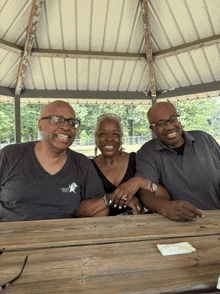 two men and a woman are posing for a picture at a picnic table in a gazebo