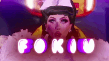 a drag queen is wearing a white hat and a white dress with purple letters behind her .