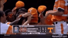 a group of people wearing orange hats with the letter t on them are standing in front of a scoreboard .