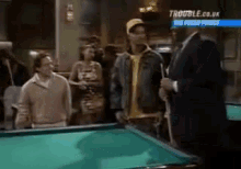 a group of people standing around a pool table with trouble.co.uk on the bottom