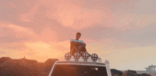 a man sits on the roof of a car with a pink sky in the background