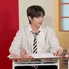 a young man in a white shirt and tie is sitting at a desk with a pen in his hand .