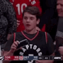 a young boy in a raptors jersey is taking a picture of a basketball game with his cell phone .