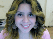 a woman wearing earrings and a pink shirt smiles at the camera