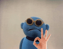 a blue stuffed animal is wearing sunglasses and making an ok sign .