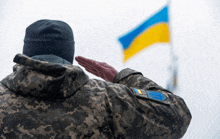 a man in a camouflage uniform salutes in front of a blue and yellow flag