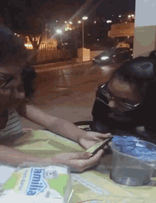 two women sitting at a table looking at a cell phone with a box of manita paper towels in the background