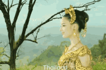 a woman in a gold dress is standing in front of a tree and the word thailand is above her