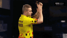 a man in a yellow shirt is holding a dart in front of a dart board with bbc america written on the bottom