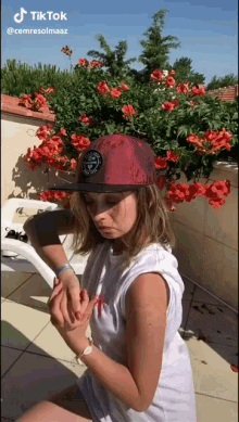 a woman wearing a red hat and a white shirt is sitting in front of a bush of red flowers ..