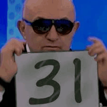 a bald man wearing sunglasses is holding a sign with the number 31 .