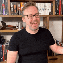 a man wearing glasses and a black shirt is laughing in front of a shelf full of books including one called emperor