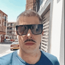 a man with a mustache wearing sunglasses and a blue shirt is taking a selfie .