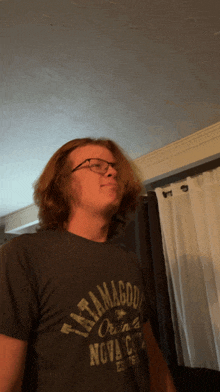 a man wearing glasses and a tatamagou t-shirt stands in a living room