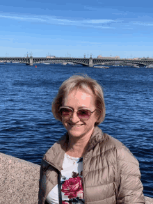 a woman wearing sunglasses and a floral shirt smiles in front of a body of water