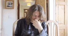 a man with long hair is sitting in a chair covering his nose with his hand