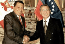 two men in suits are shaking hands in front of flags .