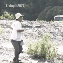 a man wearing a hat and a white shirt is standing on a rocky hillside holding a bottle of water .