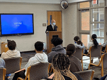 a man stands at a podium in front of a screen that says 1:07 pm on it