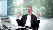 a man in a suit is sitting at a desk in front of a blackboard with chemical formulas on it