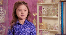 a little girl in a blue shirt is standing in front of a shelf filled with trophies and tiaras .