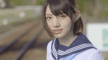 a young girl in a sailor suit is standing next to a train track and looking at the camera .