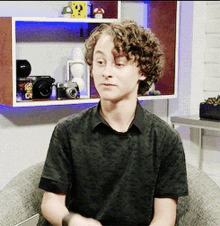 a young man with curly hair wearing a black shirt sitting in front of a shelf with mario blocks on it