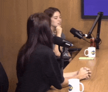 two women are sitting at a table in front of a microphone and a cup with the letter o on it .