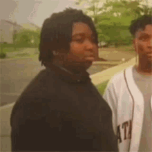 two young men are standing next to each other on a sidewalk . one of the men is wearing a baseball jersey .