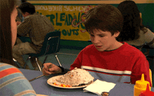 a boy sits at a table with a plate of food in front of a sign that says show your school leading outs