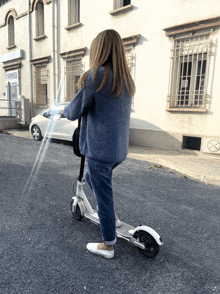 a woman in a blue sweater is riding a white scooter down a street