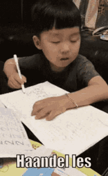 a young boy is sitting at a table writing on a piece of paper with the words haandel les written below him .