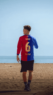 a man wearing a red and blue gavi jersey walks on a beach