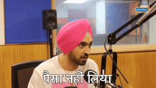 a man in a pink turban sits in front of a microphone with a radio city logo on the wall behind him