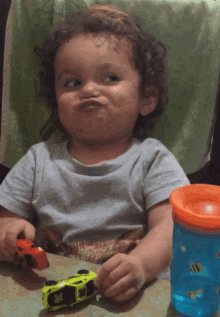 a little boy is sitting at a table playing with toy cars and a blue cup with a bee on it