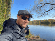 a man wearing sunglasses and a flat cap is sitting by a lake